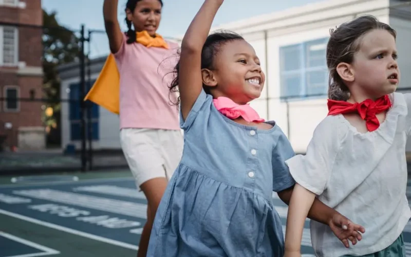 Three kids enjoying superhero play on a sunny outdoor court.