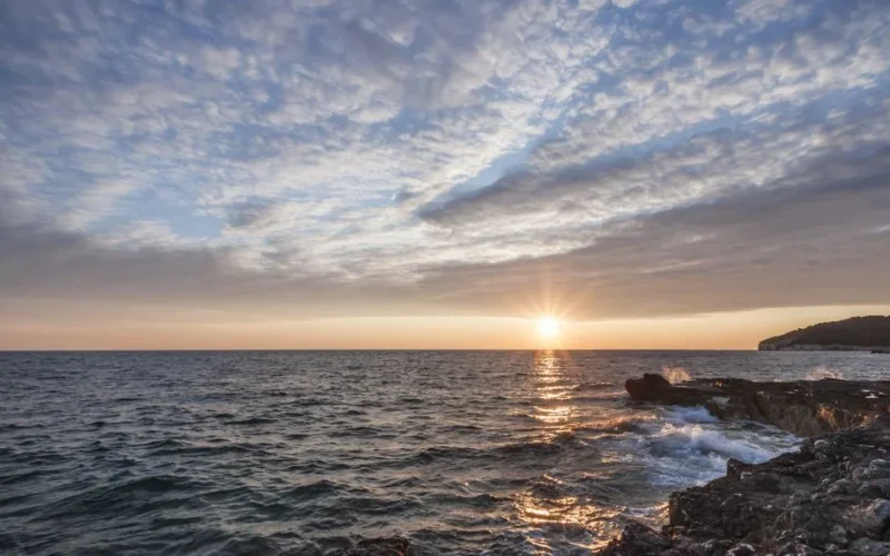 Breathtaking sunset over the ocean with dramatic clouds and rocky shoreline, perfect for serene backgrounds.