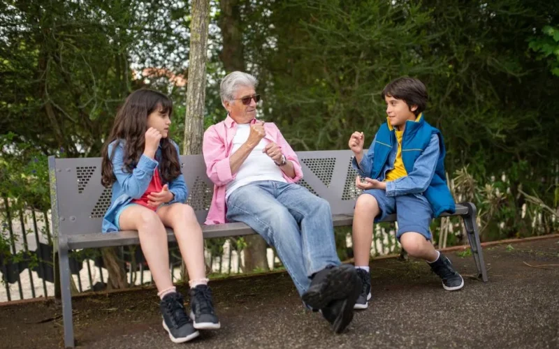 A grandfather sits with his grandchildren on a park bench, surrounded by nature, enjoying a sunny day.