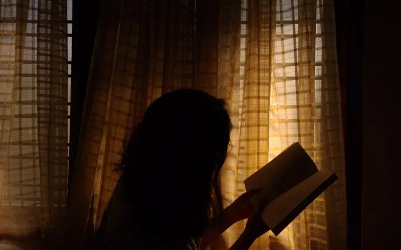 A woman enjoys a book in a tranquil room, illuminated by warm sunlight through curtains.