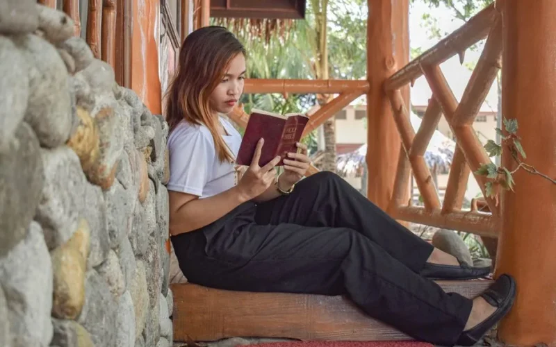 A young woman sitting on a porch reads the Bible, focused and serene in a natural outdoor setting.