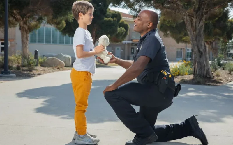 Police officer kneeling and engaging with a child holding a teddy bear in a park setting.