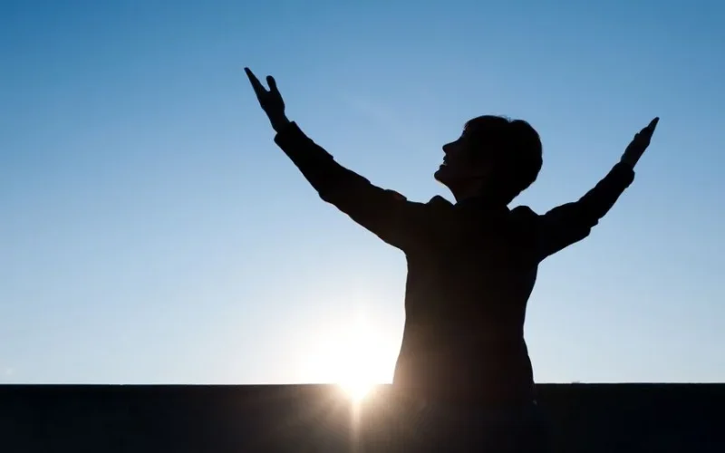 Silhouette of a person raising hands against a sunrise with a clear blue sky, exuding a sense of freedom.