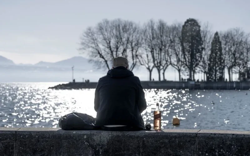 A solitary man in a jacket facing a body of water in a park, reflecting on life.