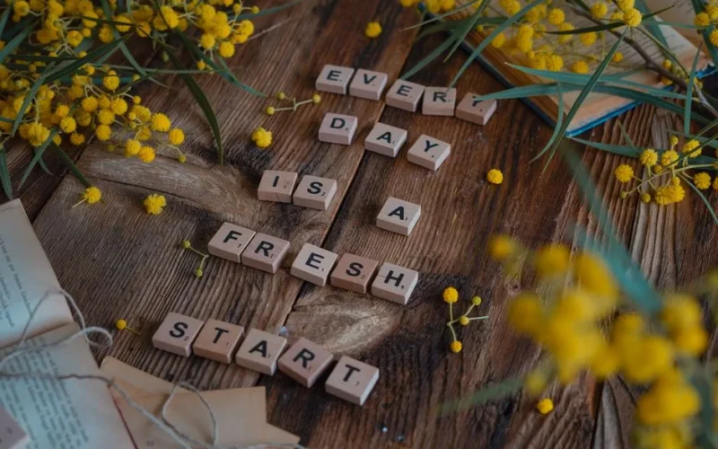Wooden tiles spell 'Every day is a fresh start' surrounded by yellow flowers on rustic table.