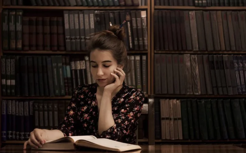 A young woman in a floral dress reads a book in a cozy library filled with vintage bookshelves.
