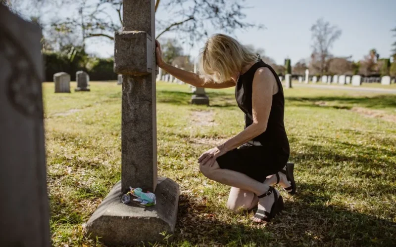 A woman in a black dress kneeling at a tombstone, expressing grief in a sunlit cemetery.