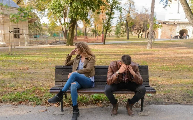 An upset couple seated on a park bench, expressing frustration during an autumn day.