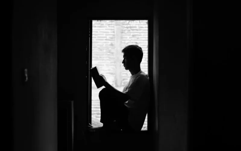 A black and white image of a person reading by a window, capturing a sense of quiet contemplation.