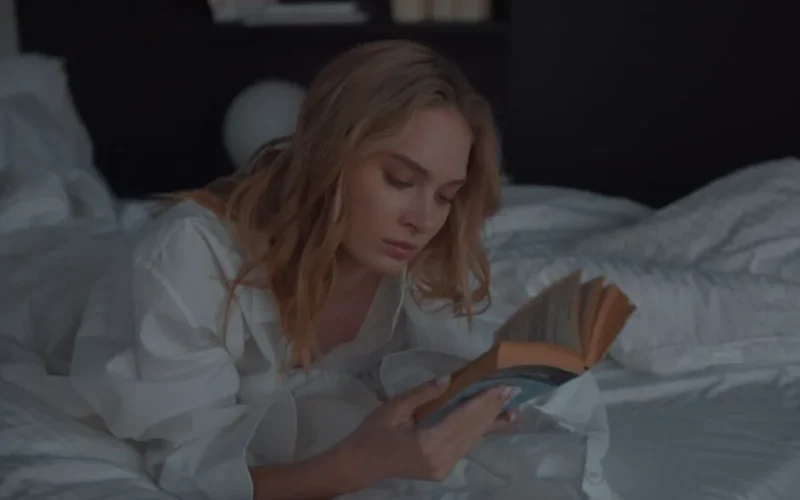 A woman lying comfortably on a bed reading a book with a calm expression in a cozy bedroom setting.