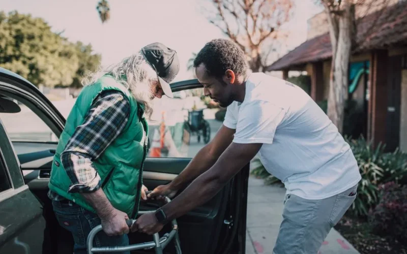 Young volunteer helping senior man with walker near a parked car, symbolizing care and support.