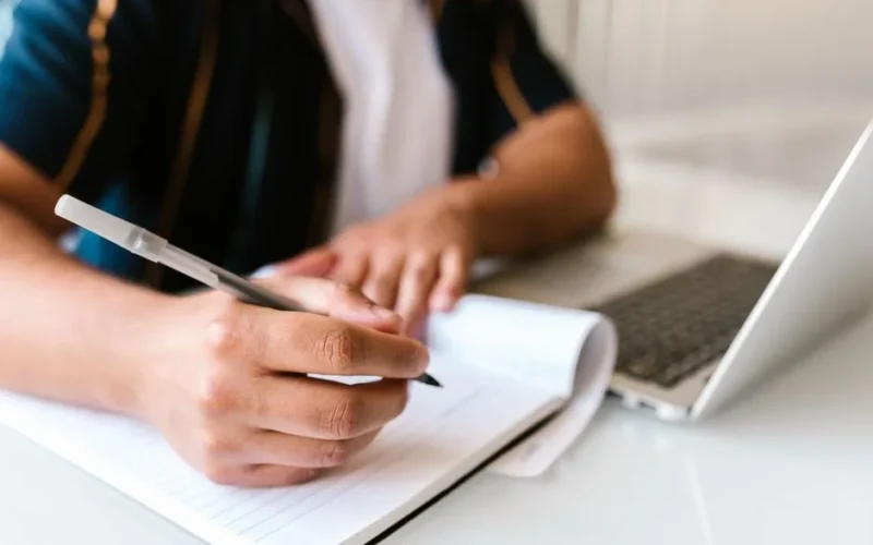 Focused image of a person writing in a notebook beside an open laptop, conveying productivity.
