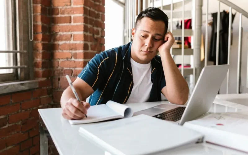 A student looks bored while studying with a laptop and notebook on a table.