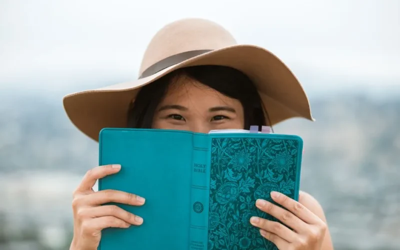 A young woman with a hat reads a blue book outdoors, showcasing a serene moment.