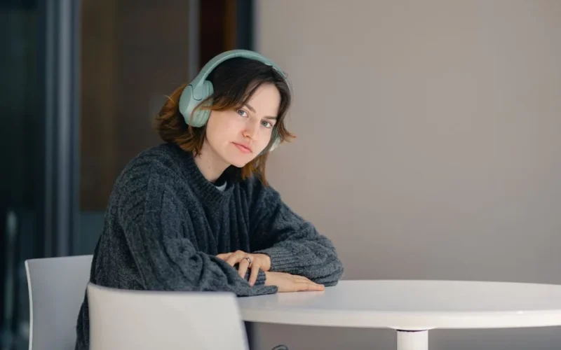 A young woman wearing headphones sits thoughtfully at a table indoors.