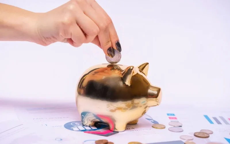 A hand places a coin into a golden piggy bank surrounded by financial documents, symbolizing savings.