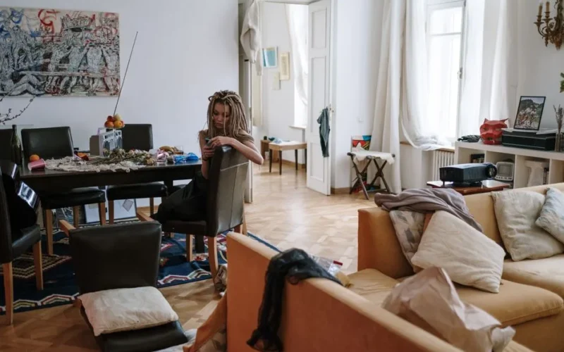 Woman with dreadlocks sitting in a cozy, cluttered room checking her smartphone.