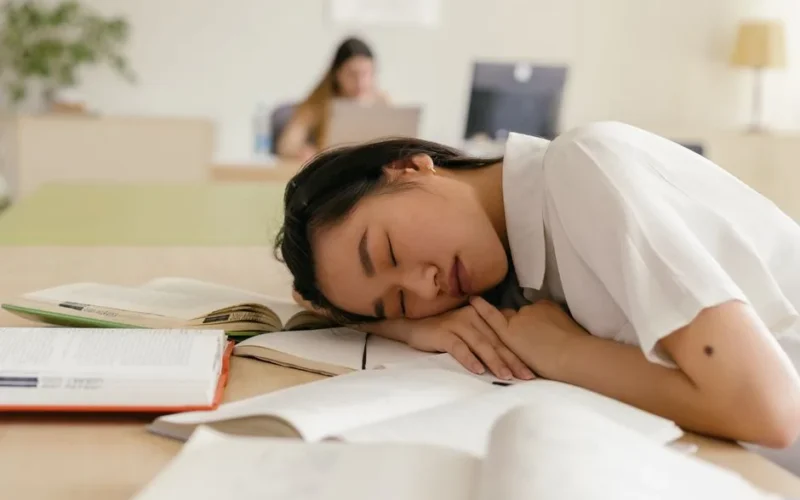 Exhausted student sleeping on a study desk covered with open books inside a quiet room.