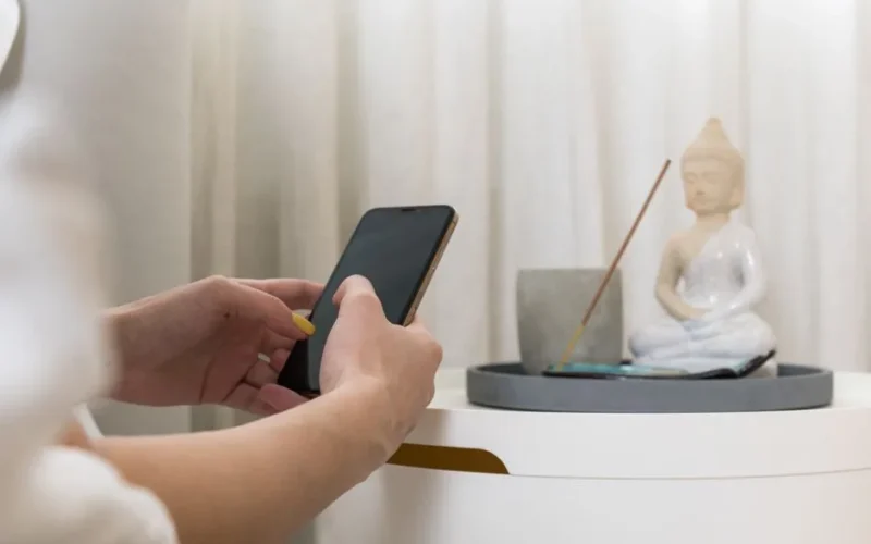 Close-up of a woman holding a smartphone near a Zen incense setup featuring a Buddha figure.