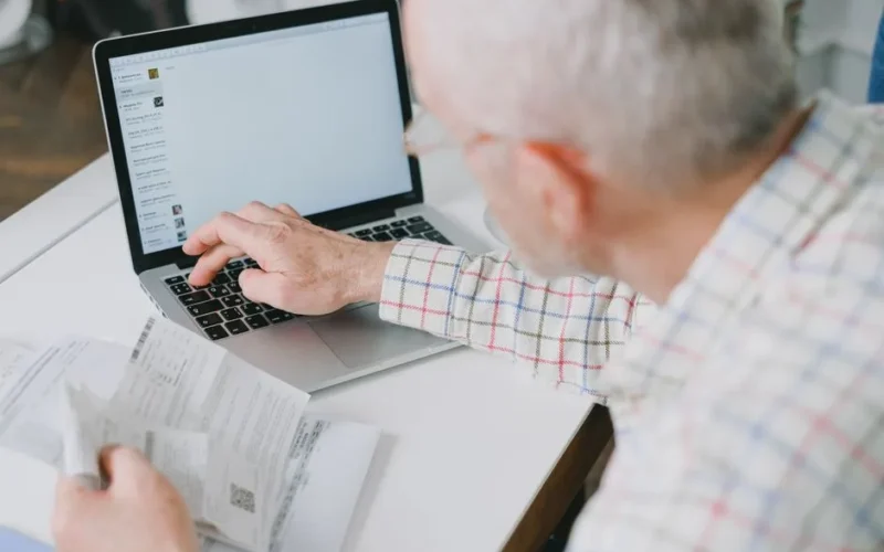 Elderly man at desk handling finances online with documents and laptop.