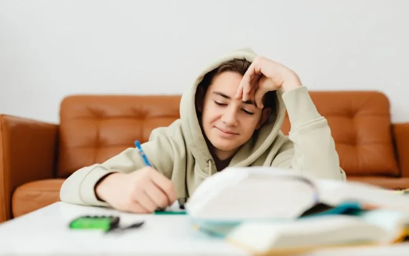 Teenage boy in hoodie smiling and writing while studying at home.