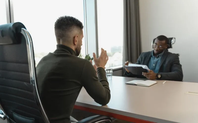 Two men in a job interview setting in a modern office with large windows.