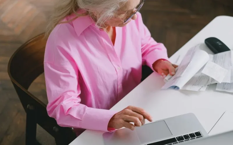 Senior woman in pink shirt using a laptop and reviewing documents at home office.