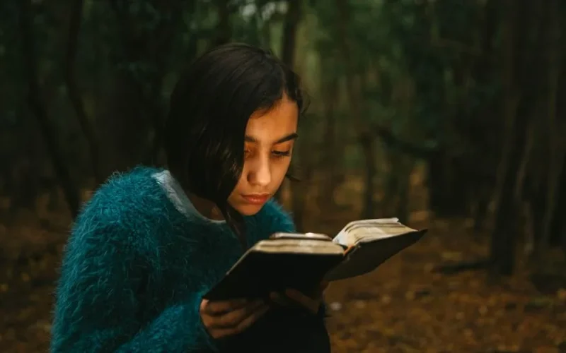 A young woman reads intently in a serene forest, wearing a cozy sweater.