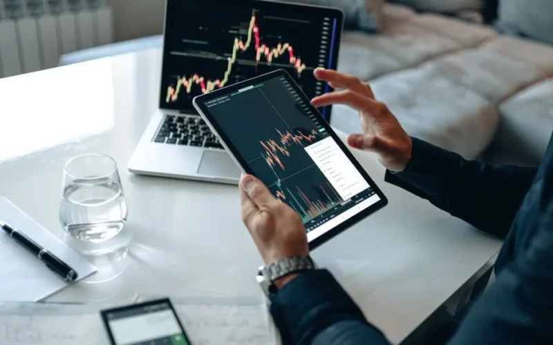 Close-up of a person using a tablet for online trading analysis with a laptop and glass of water nearby.