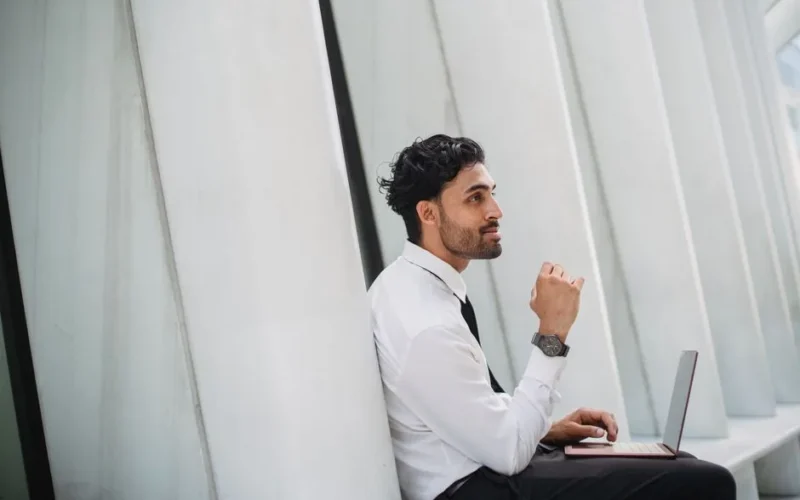 Bearded man in corporate attire using a laptop indoors with modern architecture.
