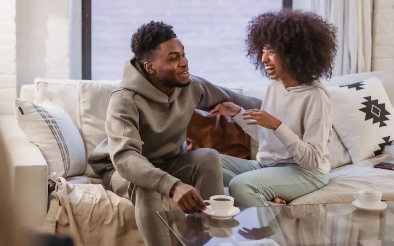 Positive African American couple talking and laughing on comfortable sofa at home in daytime