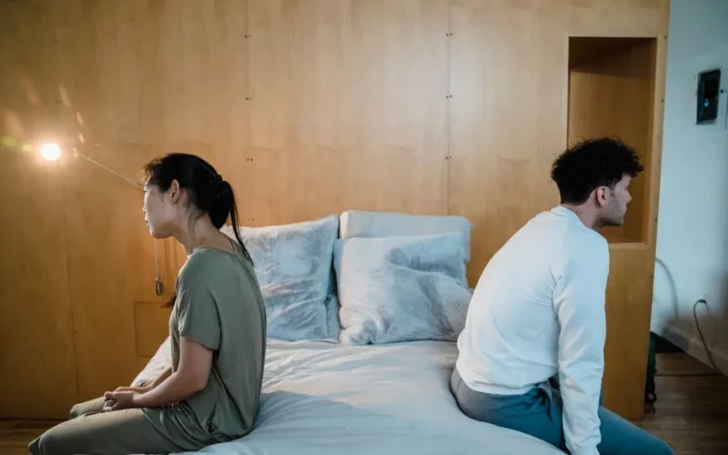 A couple sits back to back on a bed, showing tension and misunderstanding in a cozy bedroom.