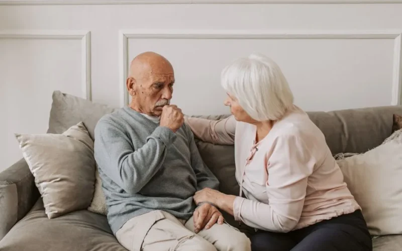 An elderly woman comforts a man coughing on a couch, showcasing care and affection.