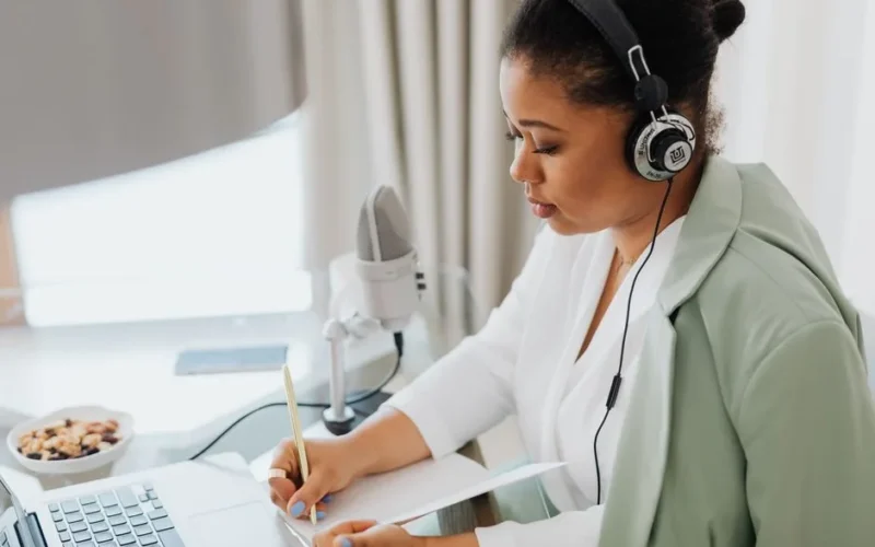 Woman wearing headphones, writing notes, and working on a laptop indoors.