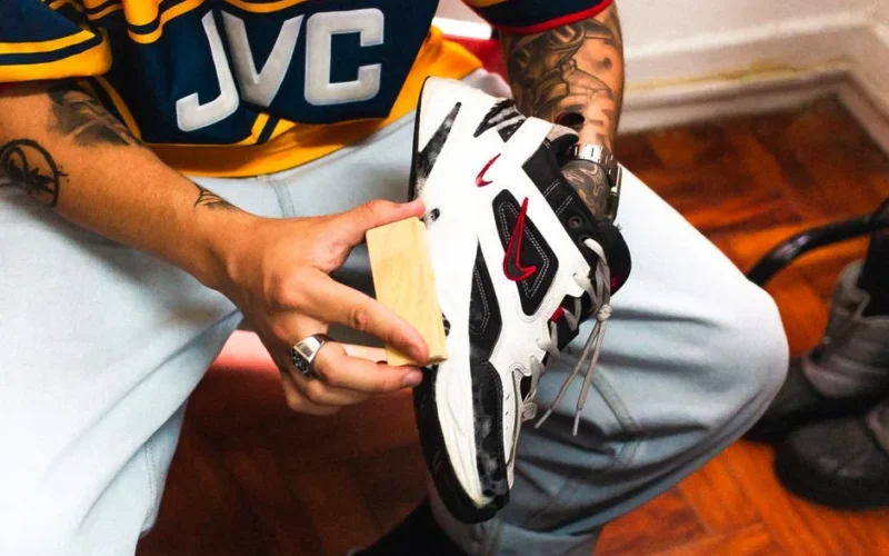 Close-up of a tattooed person cleaning a white sneaker indoors.
