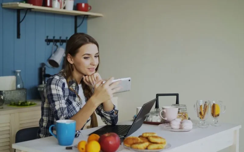 Young woman using smartphone and laptop while enjoying breakfast in cozy kitchen.
