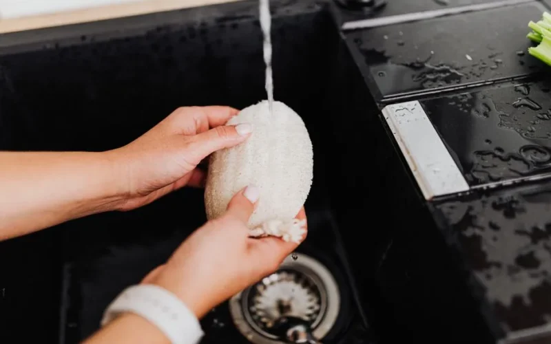 Close-up of hands cleaning a sponge under tap water in a black sink, emphasizing hygiene and kitchen cleanliness.
