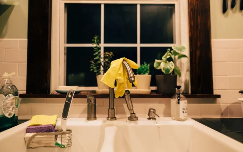 Warm lit kitchen sink at night with gloves, plants, and cleaning supplies.