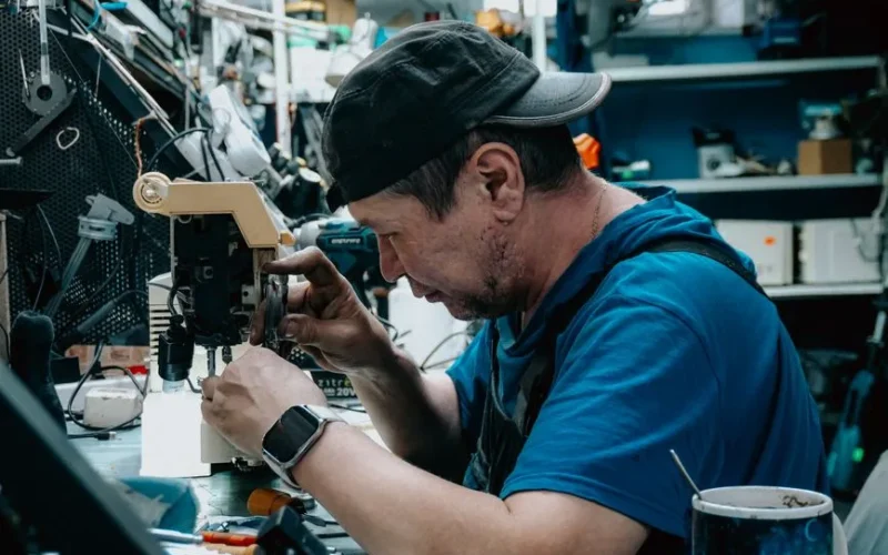 Focused man wearing cap repairs sewing machine in cluttered workshop.