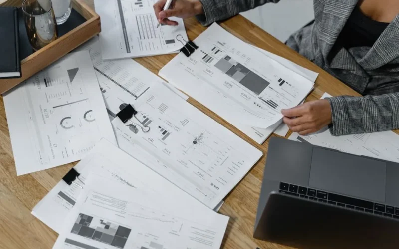 Woman analyzing financial data and reports on a wooden desk with a laptop, showcasing analytics in a workspace.