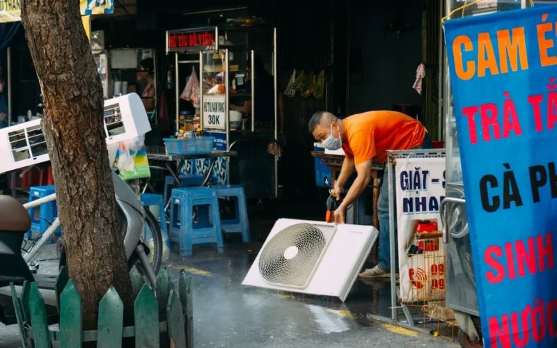 A man cleans a fan on a vibrant urban street with market stalls.