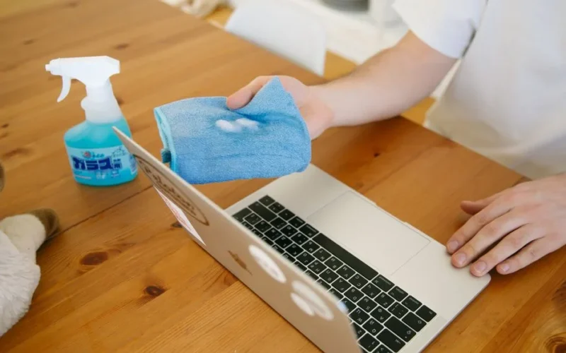Person disinfecting laptop with spray and cloth, ensuring cleanliness and hygiene indoors.