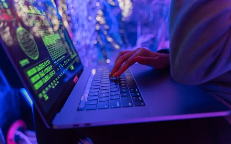 Close-up of hands typing on a laptop displaying cybersecurity graphics, illuminated by purple light.