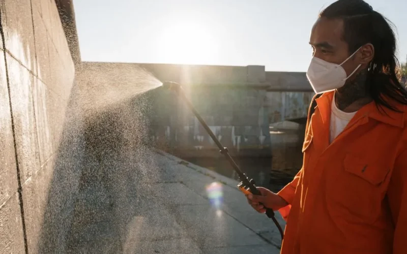 Man in orange uniform wearing facemask sprays water on wall outdoors, sunlight shining.