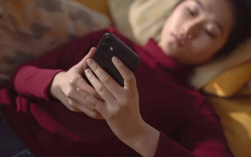 A young woman lying down, holding and using a smartphone for relaxation indoors.