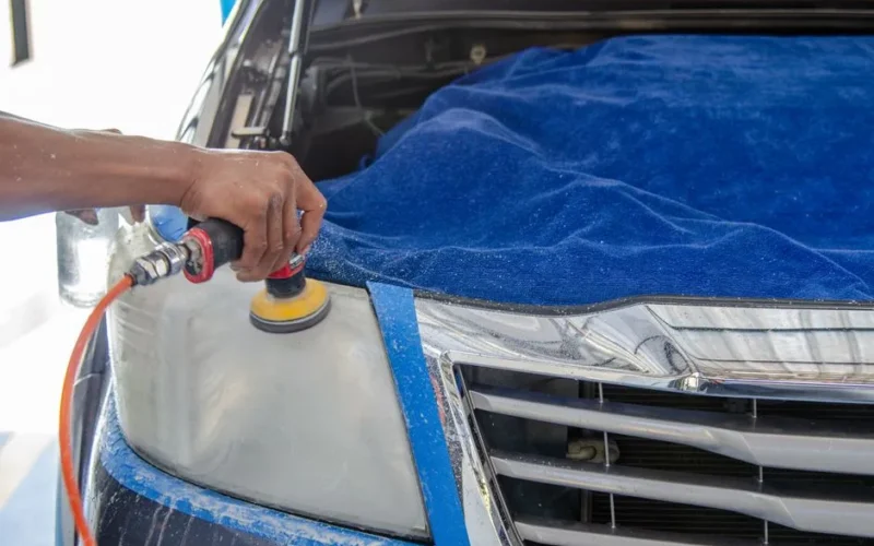 Car headlight being polished by a mechanic using an electric tool indoors.