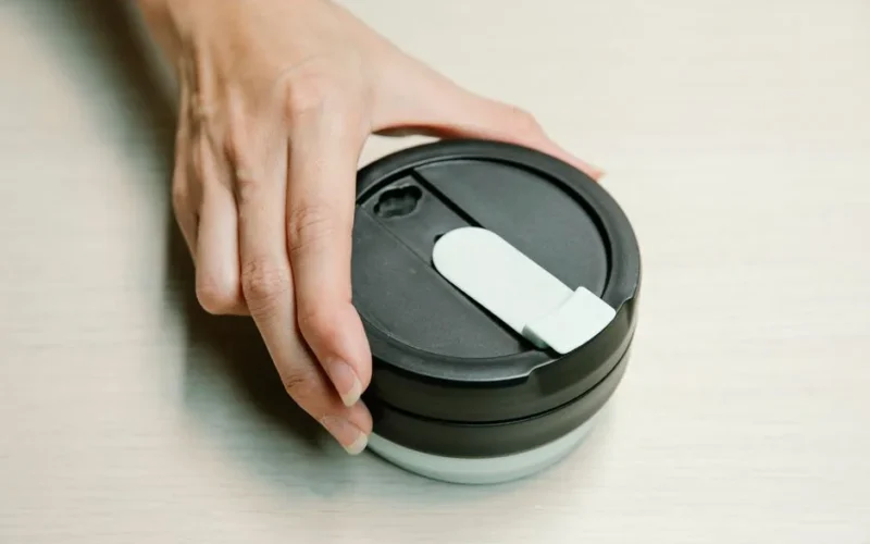 Close-up of a hand holding a black reusable coffee cup lid on a light surface.