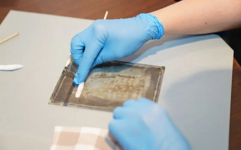 Hands in gloves carefully restoring vintage artwork on glass using cleaning tools.