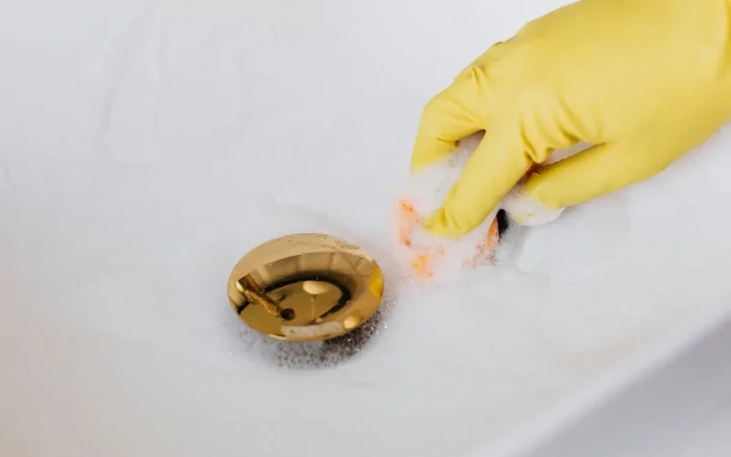From above of crop anonymous person in yellow rubber protective glove using sponge and detergent for washing white sink in bathroom