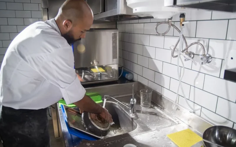 A chef cleans kitchen utensils in a stainless steel sink, maintaining hygiene standards.